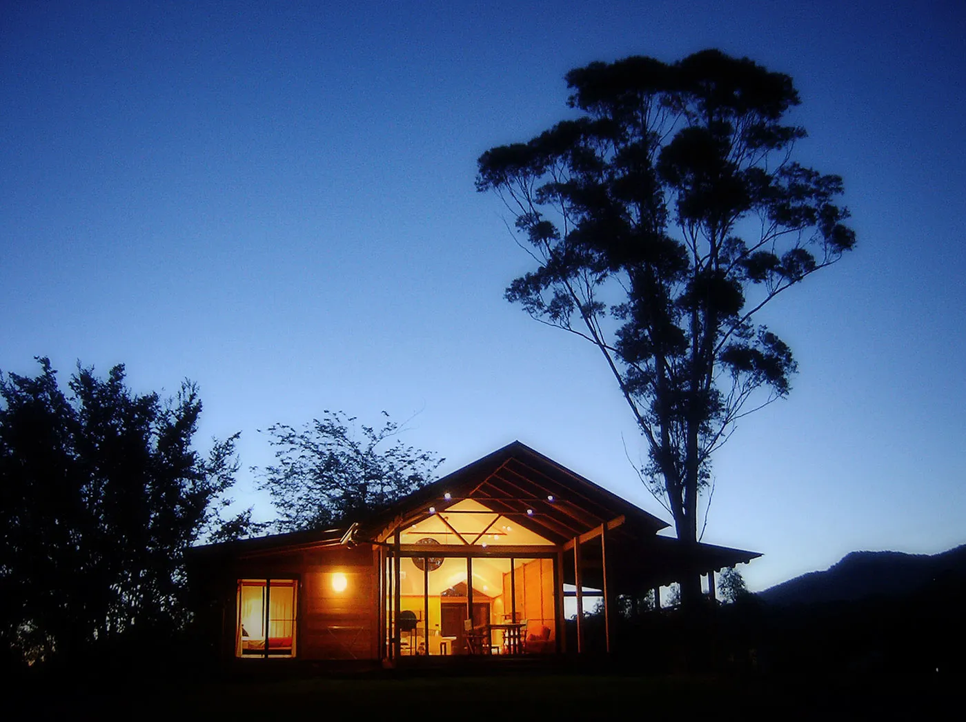 A timber chalet glowing warmly at dusk, a gum tree silhouetted against the deepening sky