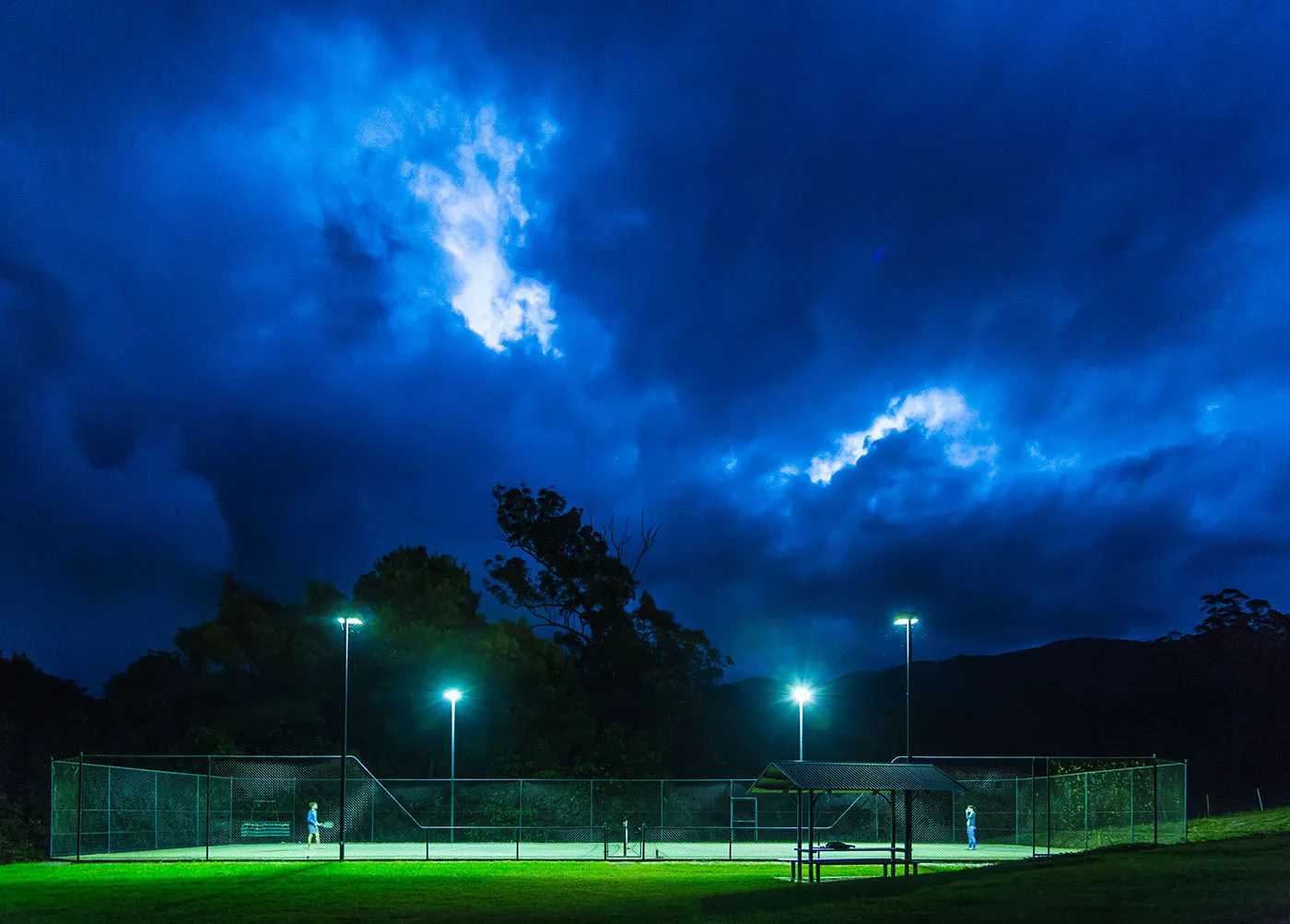 Full-size tennis court lit up for night games under floodlights, mountains silhouetted behind